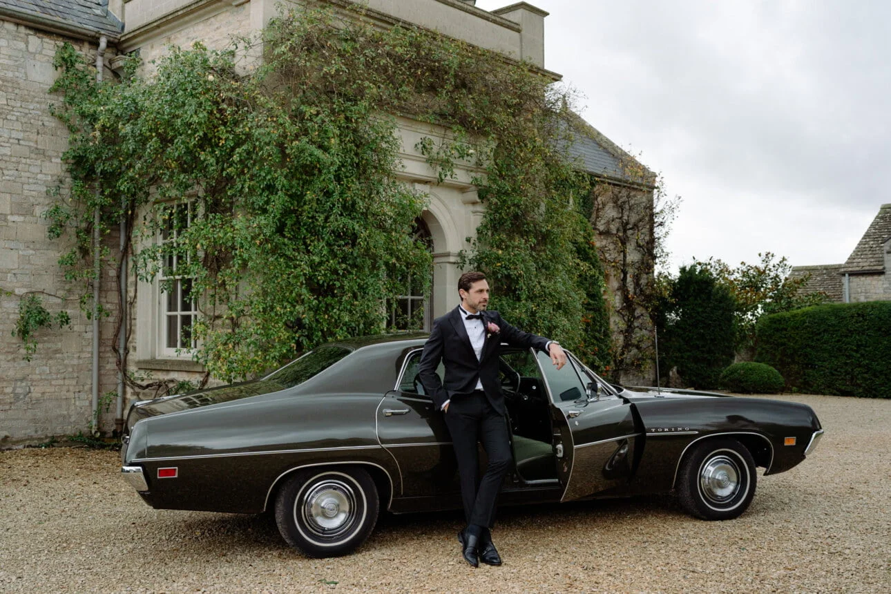 Groom wearing black tux leaning against a black car outside a Manor House